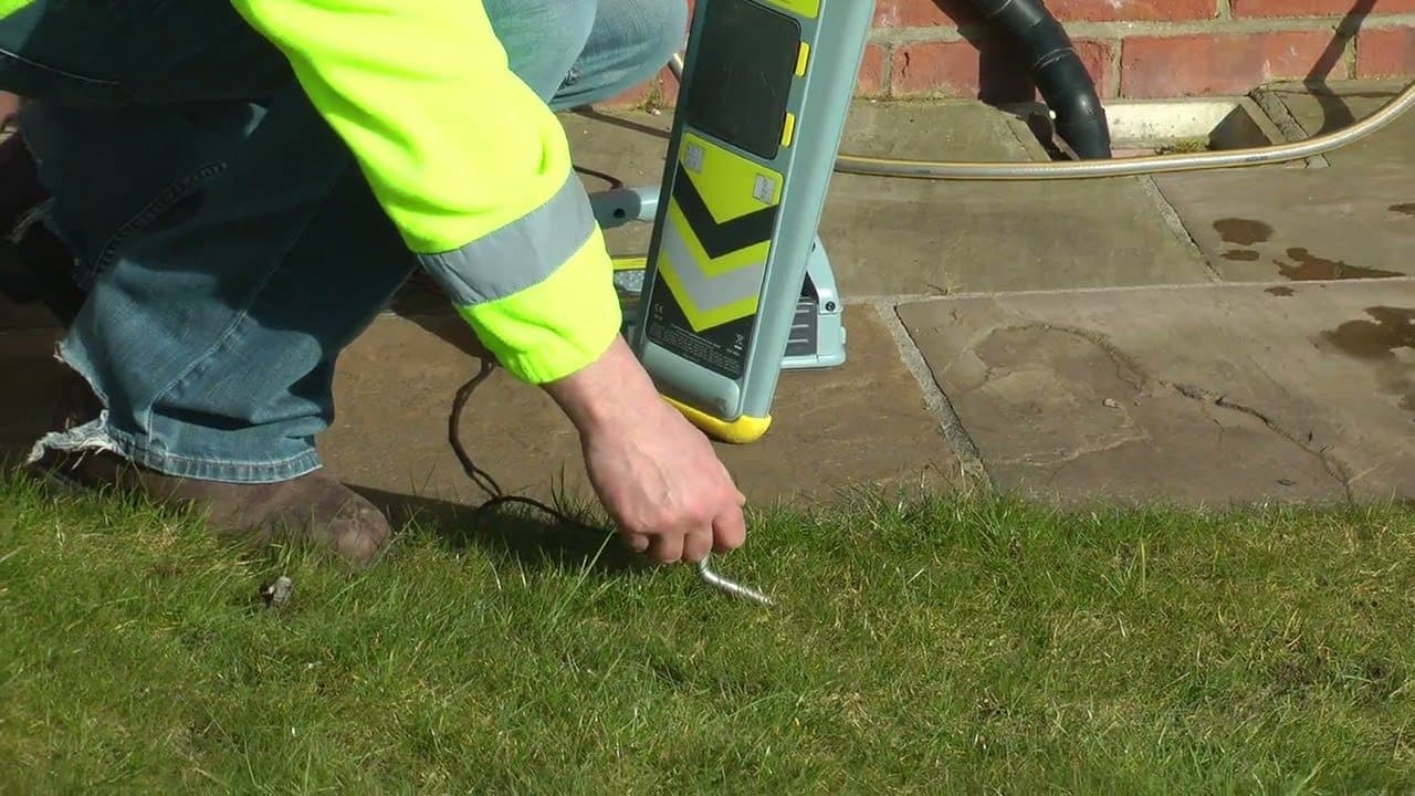 A hand pushing a Genny's earth stake into the grass beside a paved path, with a CAT standing on the paving